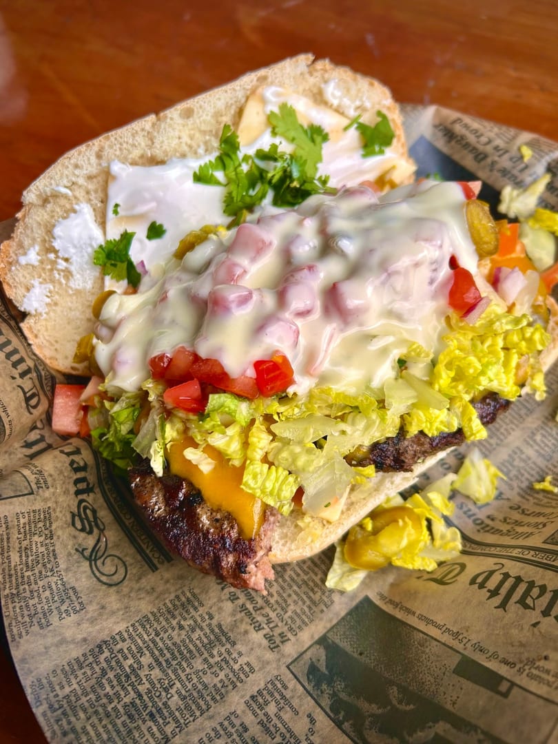 A loaded cheeseburger sub with lettuce, tomatoes, and white sauce, served on newspaper-print paper.