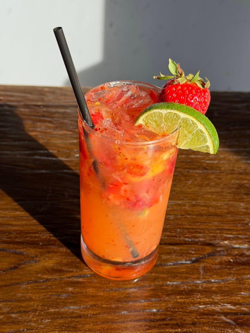 A vibrant strawberry and lime cocktail with a black straw sits on a wooden table.