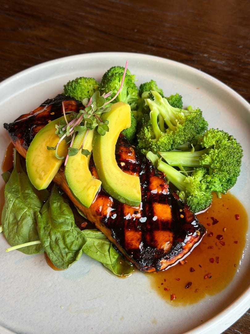 Grilled salmon with avocado slices, broccoli, spinach, and a sweet chili glaze on a white plate.