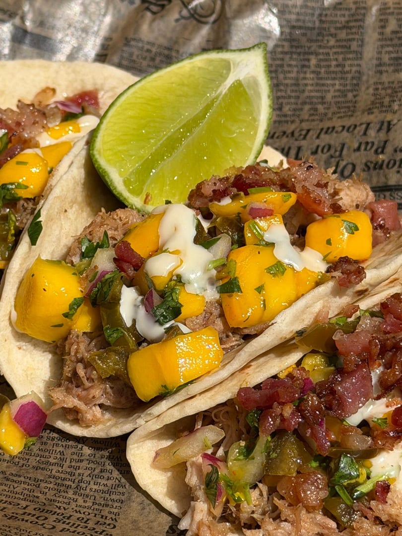 Close-up of three tacos filled with pulled pork, mango salsa, and a lime wedge.