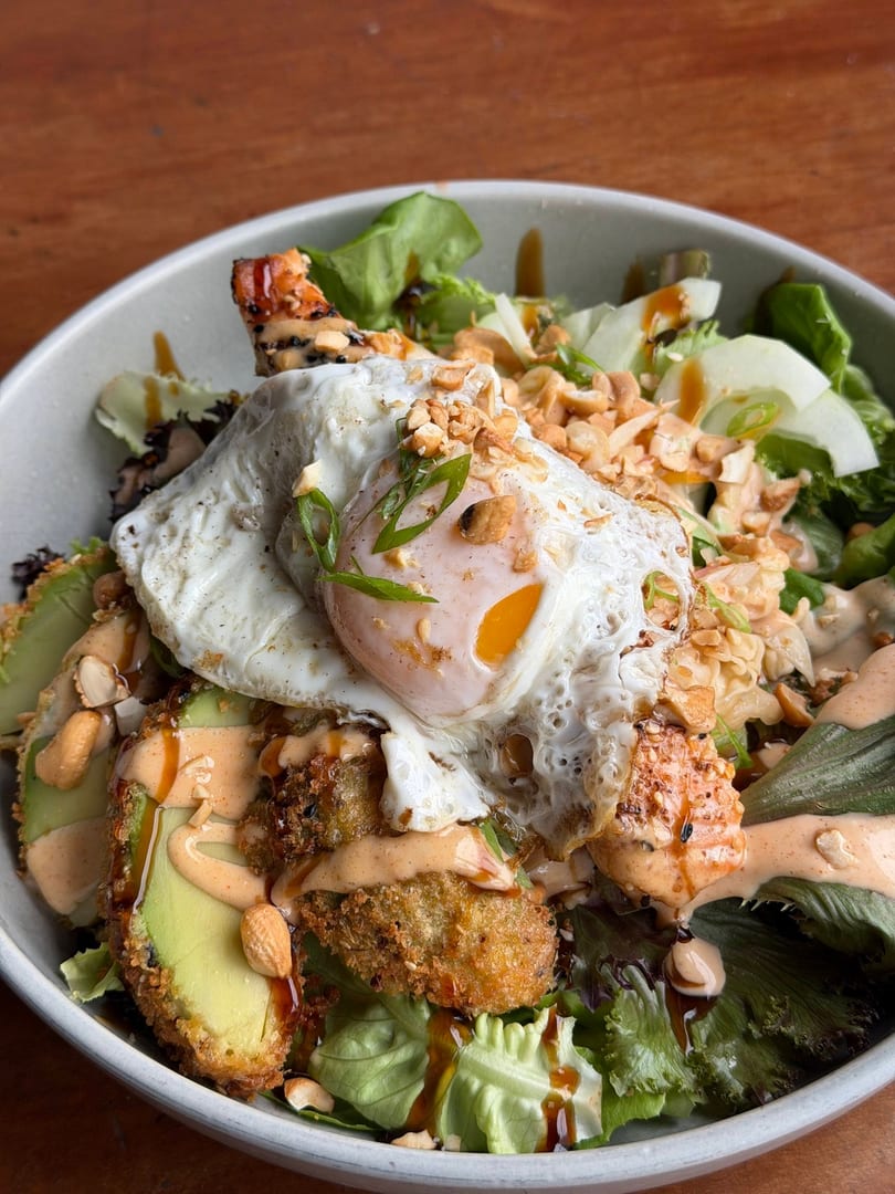 A vibrant salad bowl with fried avocado, shrimp, a fried egg, and a creamy sauce.