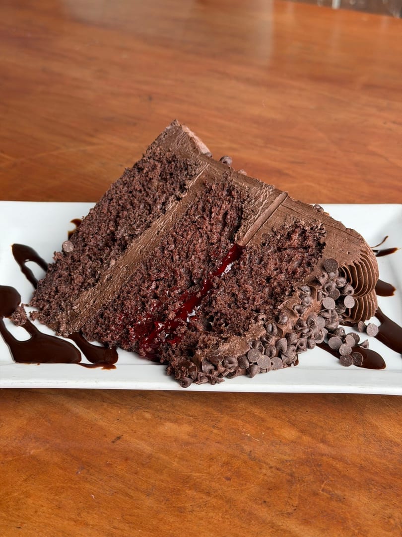 A rich chocolate cake slice with chocolate frosting, raspberry filling, and chocolate chips on a white plate.